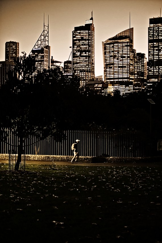 At dusk infront of a city skyline, a skateboarder rides through the park