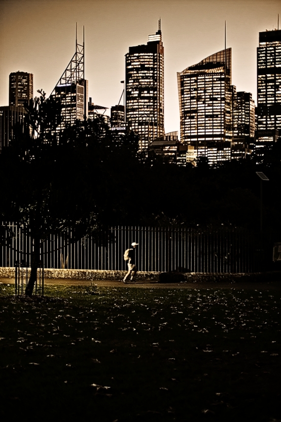At dusk infront of a city skyline, a skateboarder rides through the park